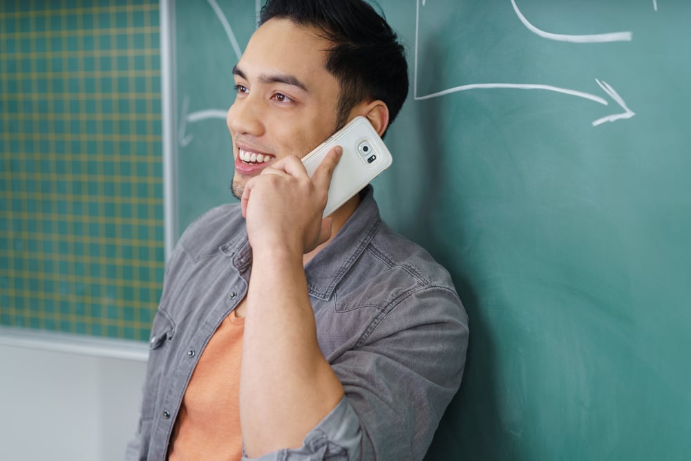 Young Asian man chatting on his mobile as he leans against a chalkboard in a college or university, close up upper body side view
