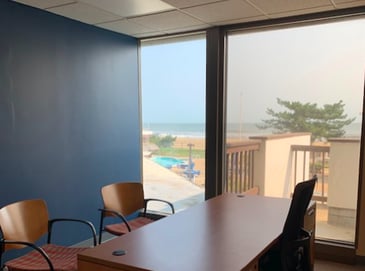 Three empty chairs around a desk in an office with a large window, where the view overlooks a beach.